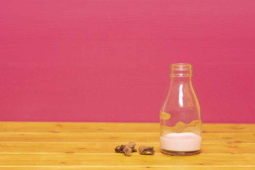 One-third pint glass milk bottle half full with strawberry milkshake and chocolate chip cookie crumbs, on a wooden table against a pink background