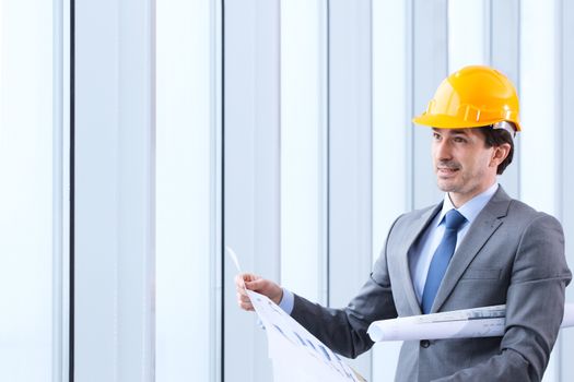 Architecture, construction business and building - smiling architect or businessman in yellow hardhat and suit with blueprint by the window