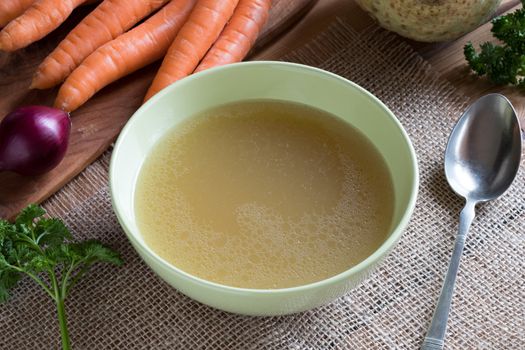 Chicken stock in a green soup bowl, with carrots, onions, parsley and celery root in the background