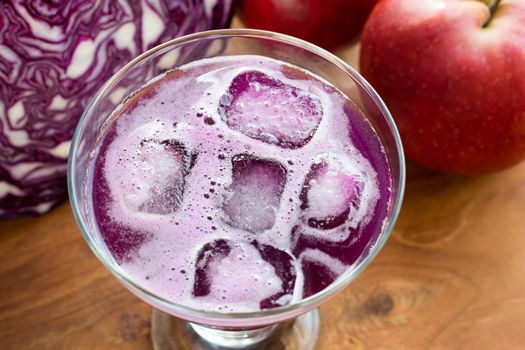 A glass of purple cabbage juice with ice cubes, with cabbage and apples in the background