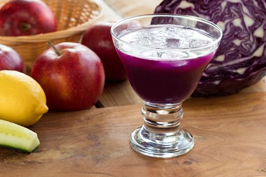 Purple cabbage juice in a glass on a wooden table, with cabbage, apples, a lemon and a cucumber in the background