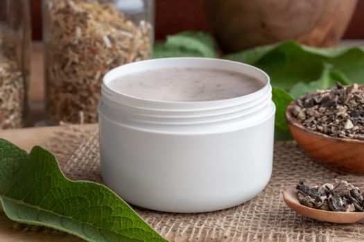 A jar of homemade comfrey ointment with fresh symphytum officinale leaves and dried root