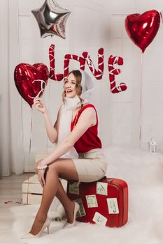 Young girl in retro style sits on a red suitcase for Valentine's Day, with inflatable balloons in the studio