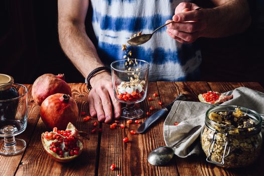 Man Puts Granola into the Glass. Series on Prepare Healthy Dessert with Pomegranate, Granola, Cream and Jam