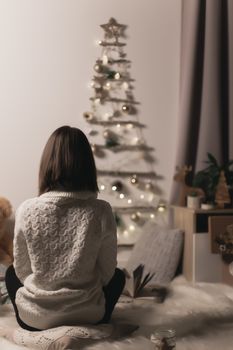 teen girl sits on the floor and looks at the Christmas tree, from the back, twilight