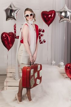 Retro girl in glasses and a scarf posing in the studio on a white background with a red suitcase and inflatable balls