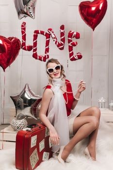 Retro girl in glasses and a scarf sits in the studio on a bench with a red suitcase and balloons