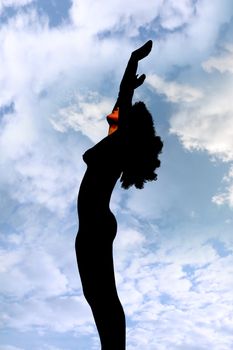 attractive silhouetted nude woman holding her hands up to the sky giving gratitude to the heavens in a yoga pose with a cloudy background