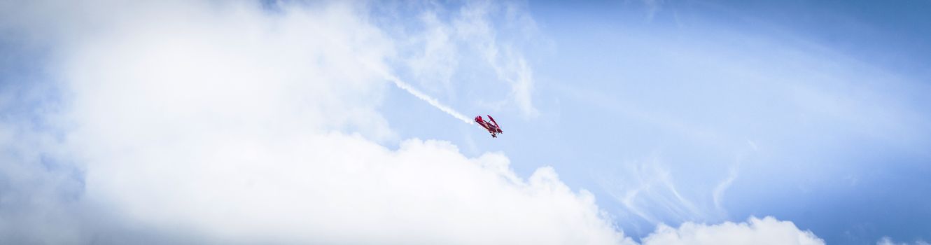 Little red airplane with propeller flying among the skies with smoke on the tail in panorama