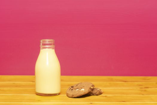 Banana milkshake in a one-third pint glass milk bottle and a chocolate chip cookie, on a wooden table against a pink painted background