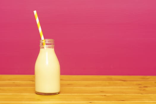 Banana milkshake with a retro paper straw in a one-third pint glass milk bottle, on a wooden table against a pink background