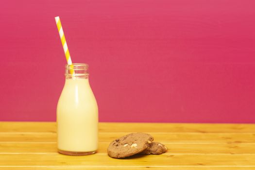 Banana milkshake with a retro paper straw in a one-third pint glass milk bottle and a chocolate chip cookie, on a wooden table against a pink background