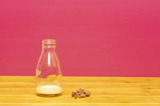 One-third pint glass milk bottle half full with banana milkshake and chocolate chip cookie crumbs, on a wooden table against a pink background