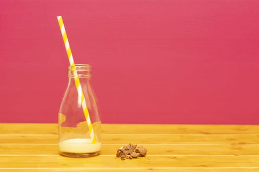 One-third pint glass milk bottle half full with banana milkshake with a retro straw and a chocolate chip cookie crumbs, on a wooden table against a pink background