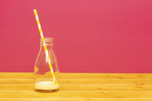 One-third pint glass milk bottle with dregs of banana milkshake with a retro paper straw, on a wooden table against a pink background