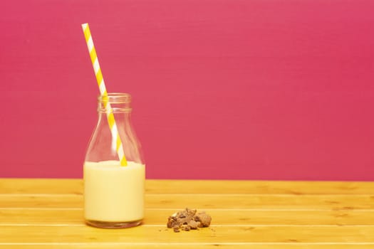 One-third pint glass milk bottle half full with banana milkshake with a retro straw and a half-eaten chocolate chip cookie, on a wooden table against a pink background