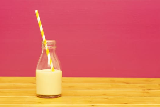 One-third pint glass milk bottle half full with banana milkshake with a retro paper straw, on a wooden table against a pink background