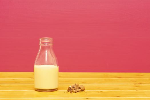 One-third pint glass milk bottle half full with banana milkshake and a half-eaten chocolate chip cookie, on a wooden table against a pink background