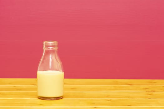 One-third pint glass milk bottle half full with banana milkshake, on a wooden table against a pink painted background