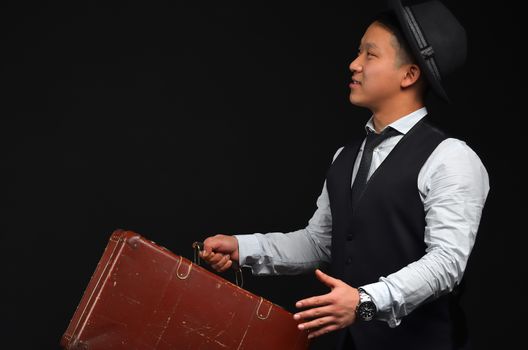 Asian young man in a hat with a brown suitcase, and raises his arms looking sideways