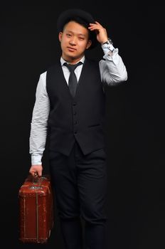 The photo of an Asian man in full height, he carries a brown suitcase and touches his hat