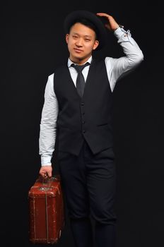 A photo of an Asian man in full height, he carries a brown suitcase and raises his hat by congratulating