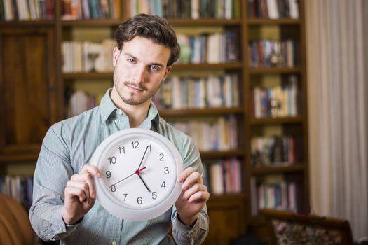 Young casual man holding clock and looking pressed with time limit posing inside at home