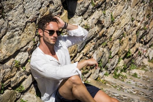 Side view of stylish man in sunglasses sittingon stone stairs and relaxing. Ocean on background.
