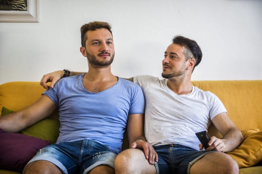 Two gay men in casual outfits sitting on sofa embracing and watching TV at home