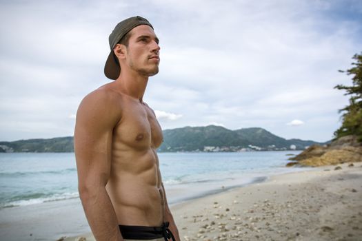 Attractive shirtless athletic young man on beach by the sea, with baseball cap and sunglasses, looking away