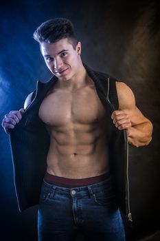 Handsome young muscular man shirtless wearing jeans and sleeveless vest, on black background in studio shot