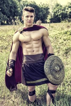 Young handsome muscular man posing in roman or spartan gladiator costume with shield and sword on field.