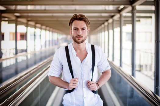 Handsome young man walking in city with backpack on shoulders, wearing white shirt