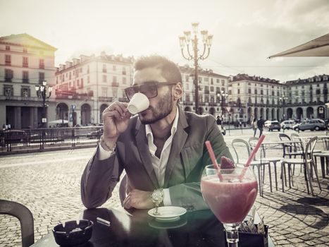 Handsome young man drinking espresso coffee, wearing elegant coat posing at table outside on urban background.