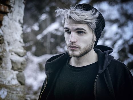 Portrait of young man in hoodie posing outdoor in winter setting with snow all around, looking away to a side.