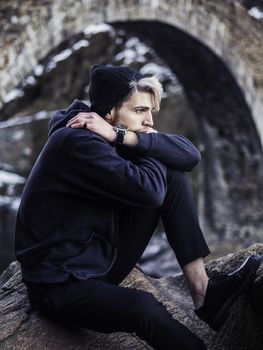 Portrait of young man in hoodie posing outdoor in winter setting with snow all around, looking away to a side.