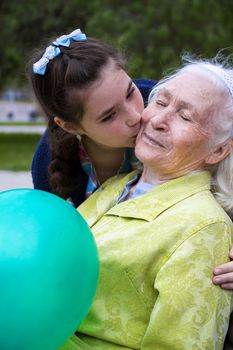 Pretty girl hugs and kisses his beautiful gray-haired grandmother on the cheek. A pleasant elderly woman sits on a park bench holding a balloon and smiles shyly.