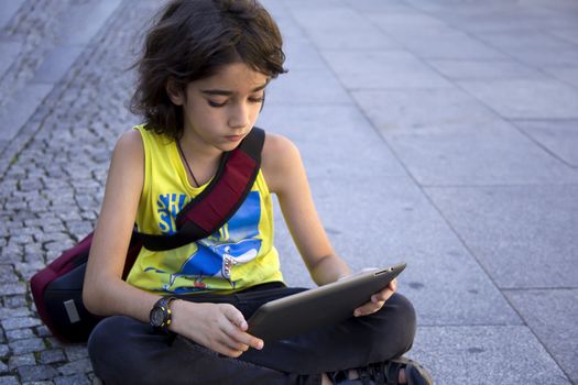Teenage boy with long brown hair sitting outside on concrete using a tablet computer, looking down while sitting with crossed legs.