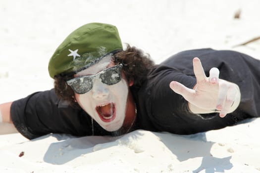A young guy in sunglasses is lying on a white sandy beach and fooling around, smearing his face in the sand and shouting joyfully. The concept of happiness, relaxation, no problem, everything is fine.