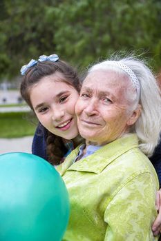 Beautiful girl hugging his beautiful gray-haired grandmother. A pleasant elderly woman sits on a park bench holding a balloon and they both smile happily.