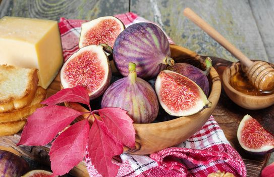 Beautiful autumn still life: ripe figs in a wooden bowl, red raspberry, toasts, cheese, honey and a checkered napkin on old cutting board as well as autumn leaves on the old wooden table