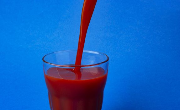 Tomato juice pouring into glass, isolated on blue background, with copy space, healthy drink concept, close-up