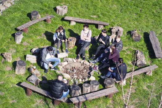 A group of people are sitting by the fire. in a camp with a guitar, tent, blanket, backpacks, sitting near the fire place in a camp.Spring warm day.