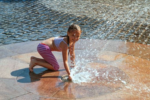 Girl on a sunny summer day are poured water from a fountain.Girl happily in shallow clean water on of city fountain on warm bright summer day.
