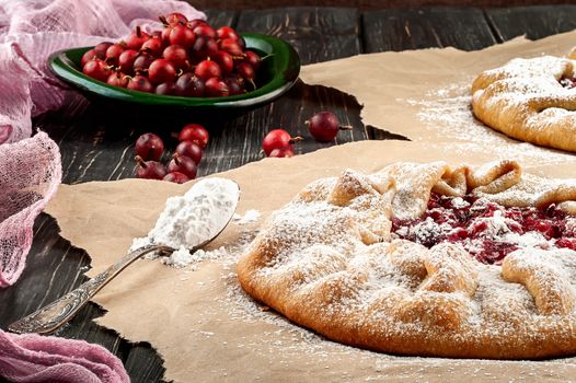 Open pies with gooseberries on a wooden table. Spoon with powdered sugar nearby. Gooseberry on a green plate in the background.