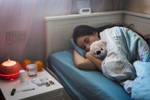 The young girl has a cold, lies in bed at home, feels unwell, takes medicines and vitamins, sleeps with a toy bear.