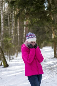 Portrait of a teenage girl in a bright pink winter jacket and knitted hat, which covers her face with a scarf. She stay in the winter snowy forest.