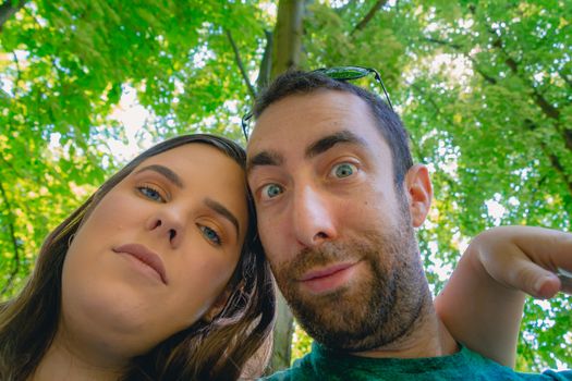 Happy young couple taking a selfie with a smartphone or camera at the park. They looking down while making silly and funny faces.