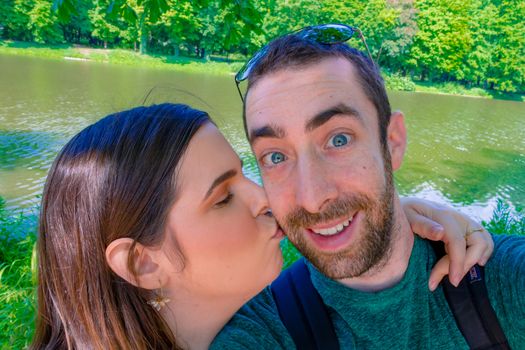 Happy young couple taking a selfie with smartphone or camera at the park. The man smiling while the woman kisses him on the cheek.