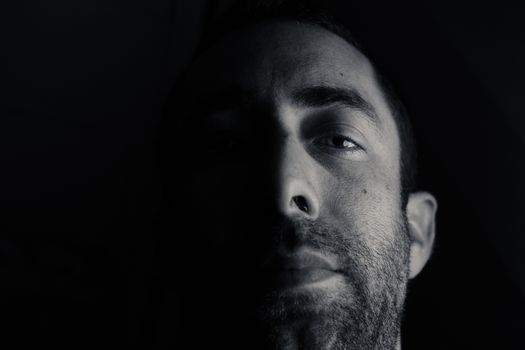 Dramatic portrait of a young man half expressionless face in deep shadow looking at camera isolated on black background.